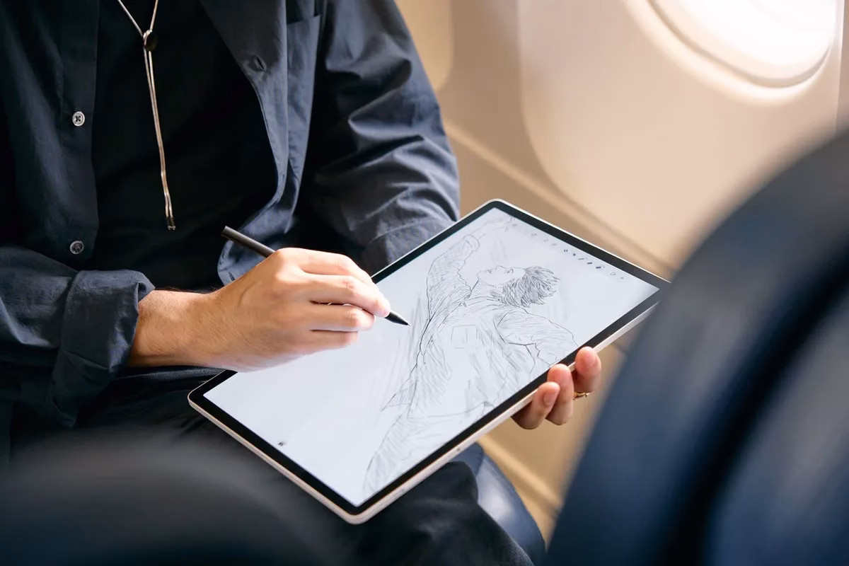 飛行機内でタブレットを使って絵を描いている人物の写真。男性がタブレットを手に持ち、ペンでスケッチを描いている様子が写っています。タブレットの画面には人物のデッサンが描かれています。