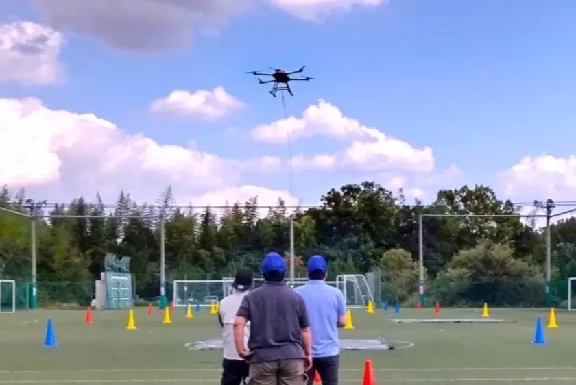 空に浮かぶドローンと、それを見守る人々の様子を捉えた写真です。ドローンはケーブルで繋がっており、操作されているようです。人々の背後には、コーンが並んだグラウンドがあり、ドローンの飛行訓練が行われている様子が伺えます。