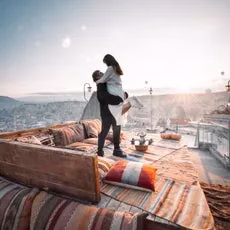 男性が女性を抱き上げている様子。背景には街並みと夕焼けが見える。