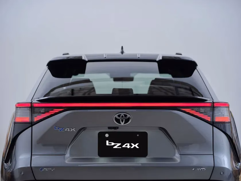 灰色のトヨタbZ4Xの車両後部を捉えた画像です。特徴的な一文字の赤いLEDテールランプが点灯しており、ナンバープレートと車体には「bZ4X」のロゴが見えます。BEV（電気自動車）でAWDモデルであることが確認できます。