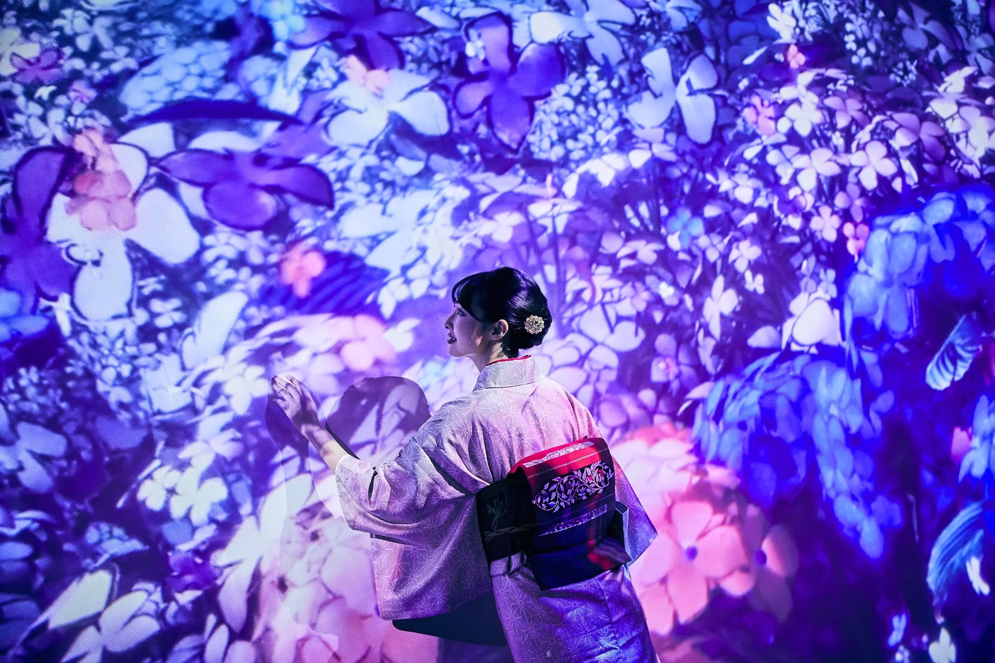 着物を着た女性が、花が咲き乱れる映像が映し出された空間に立っています。女性は右手を伸ばし、花に触れようとしているかのように見えます。