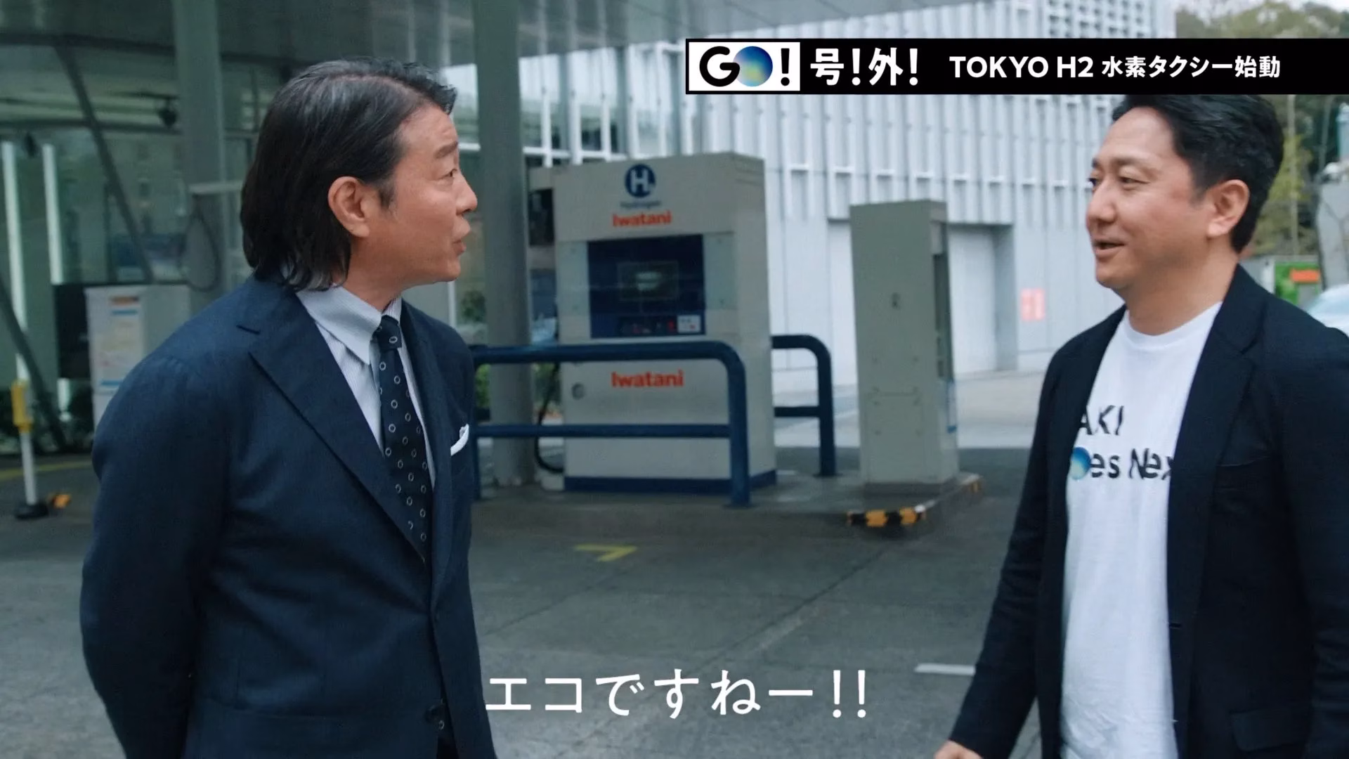水素ステーションの前で話す2人の男性。1人はスーツ姿、もう1人はTシャツにジャケット姿。背景には「TOKYO H2 水素タクシー始動」の文字。