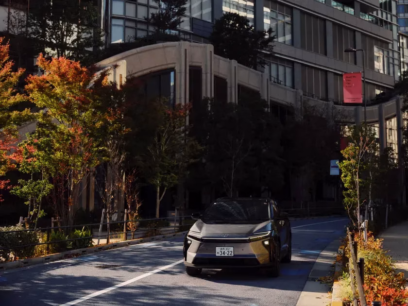 秋の都市景観の中、現代的なデザインの黒いSUVが道路を走行しています。周囲には紅葉した木々やモダンなビルが立ち並び、日本の秋の街並みを表現しています。