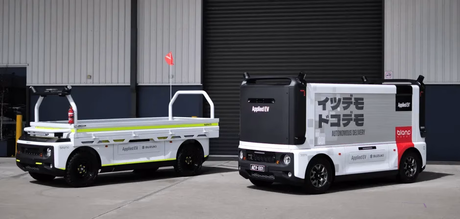 2台の白い電気自動車が並んで駐車されている写真。左側の車両は荷台付きで、右側の車両は箱型の構造をしている。どちらの車両にも「Applied EV」のロゴと「SUZUKI」の文字が見える。