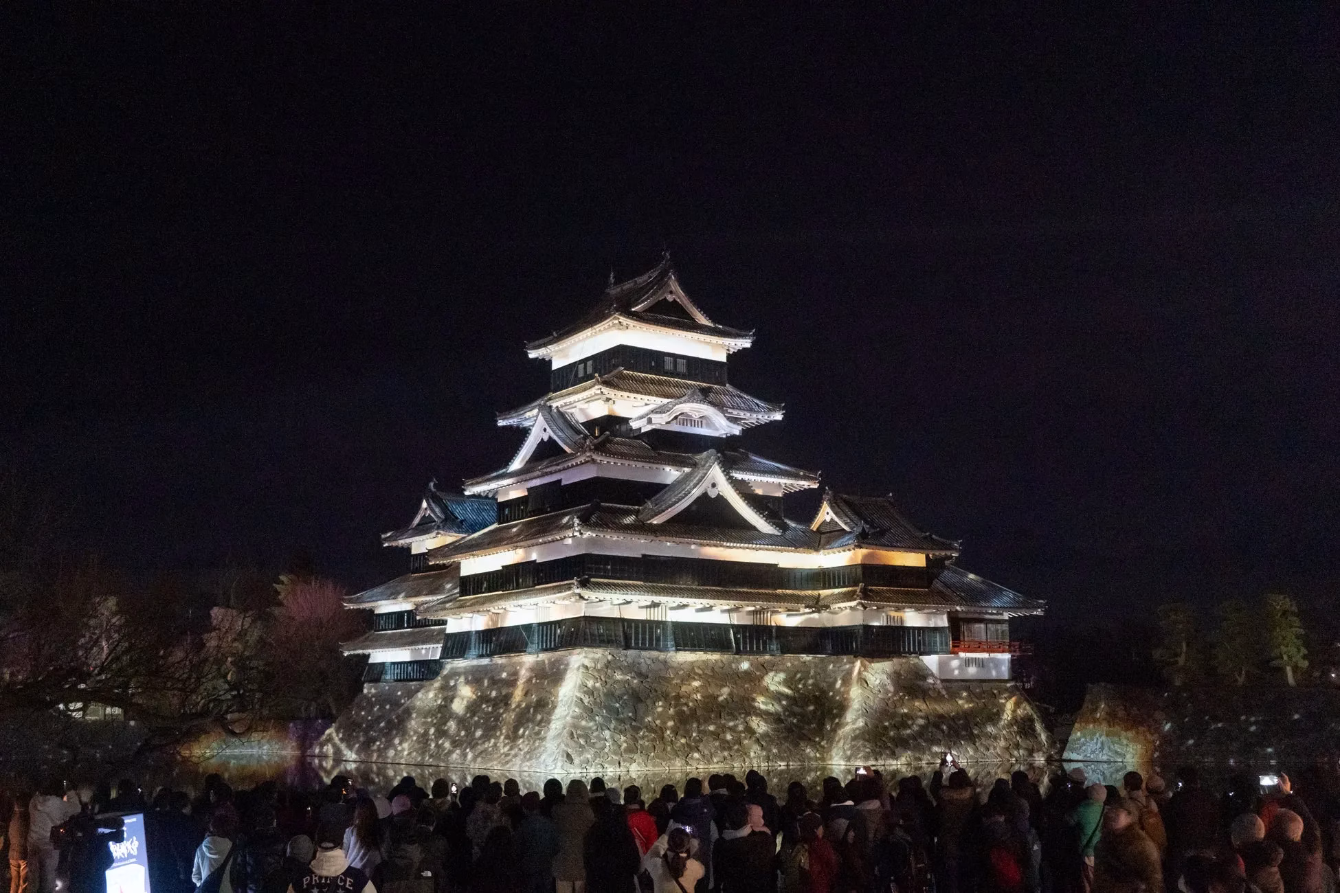 夜の松本城の画像。ライトアップされた城と、それを見物する人々の姿が写っている。
