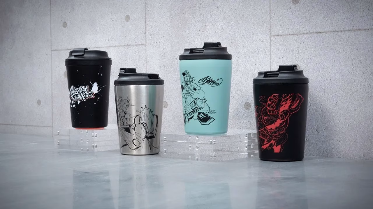 4つのタンブラーが並んでいる画像。タンブラーにはそれぞれ異なるイラストが描かれており、黒、シルバー、水色、黒の4色で、すべて黒い蓋が付いている。タンブラーは透明な台の上に置かれ、背景はコンクリートのような質感の壁。