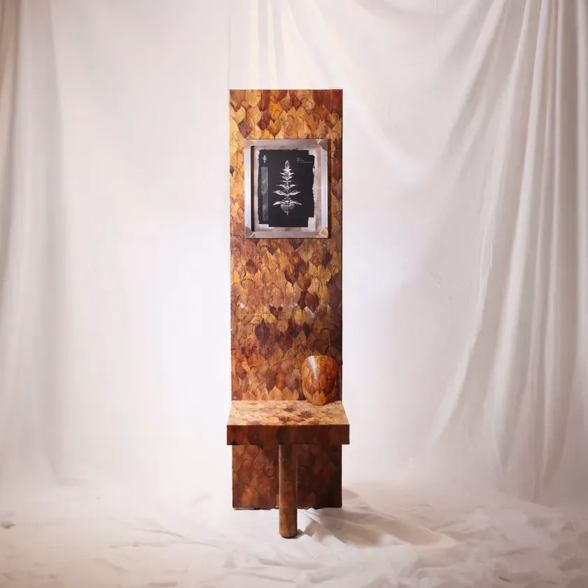 茶色を基調とした、葉の模様が施された背もたれと、その下に小さな台座がついた椅子のようなオブジェ。正面には植物の絵が飾られており、台座には壺のようなものが置かれている。背景は白い布で覆われている。