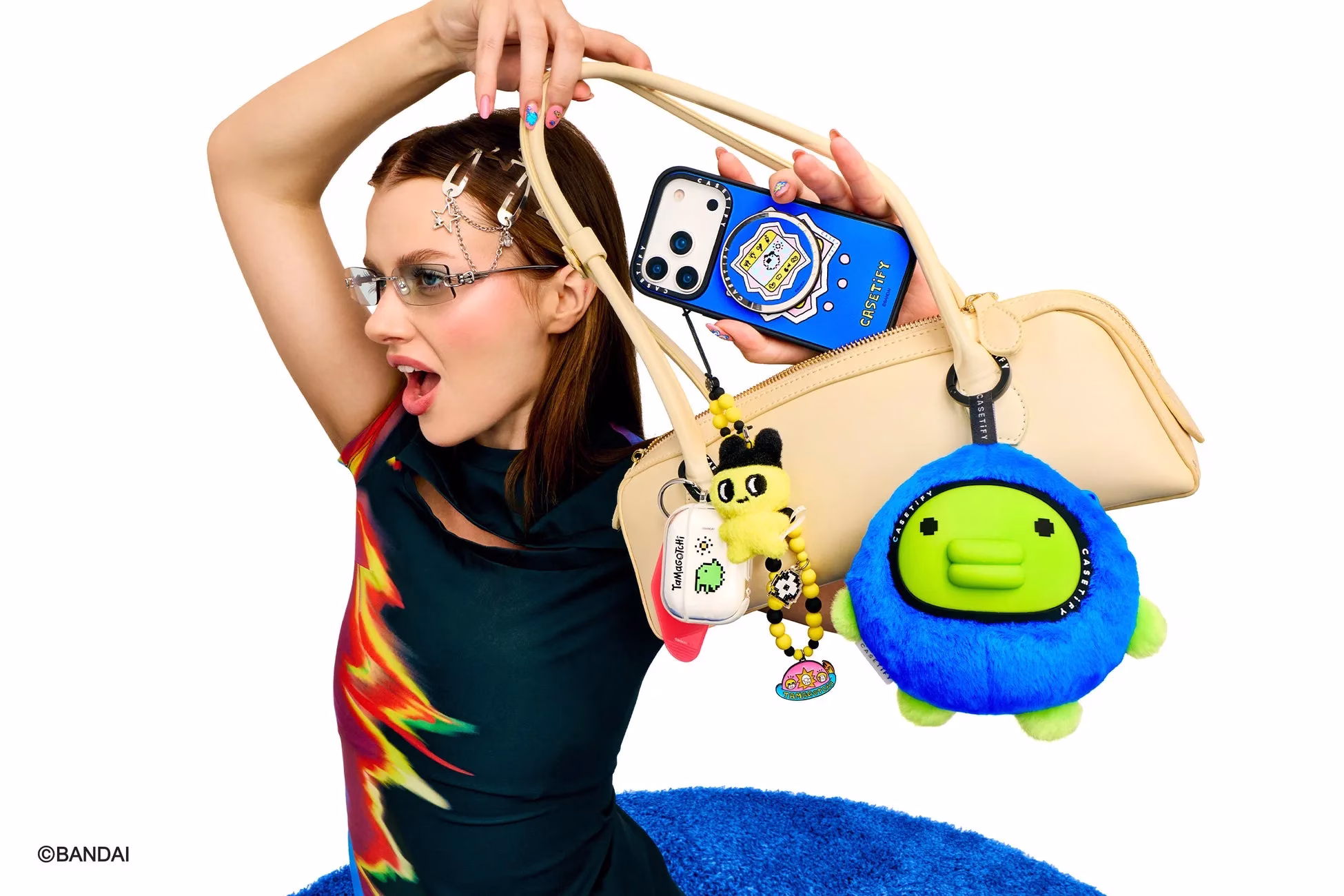 女性がバッグを持ち、スマートフォンケースやアクセサリーを見せている写真。彼女はメガネをかけ、驚いたような表情をしている。バッグにはTamagotchiのアクセサリーやCASETiFYのケースが付いている。