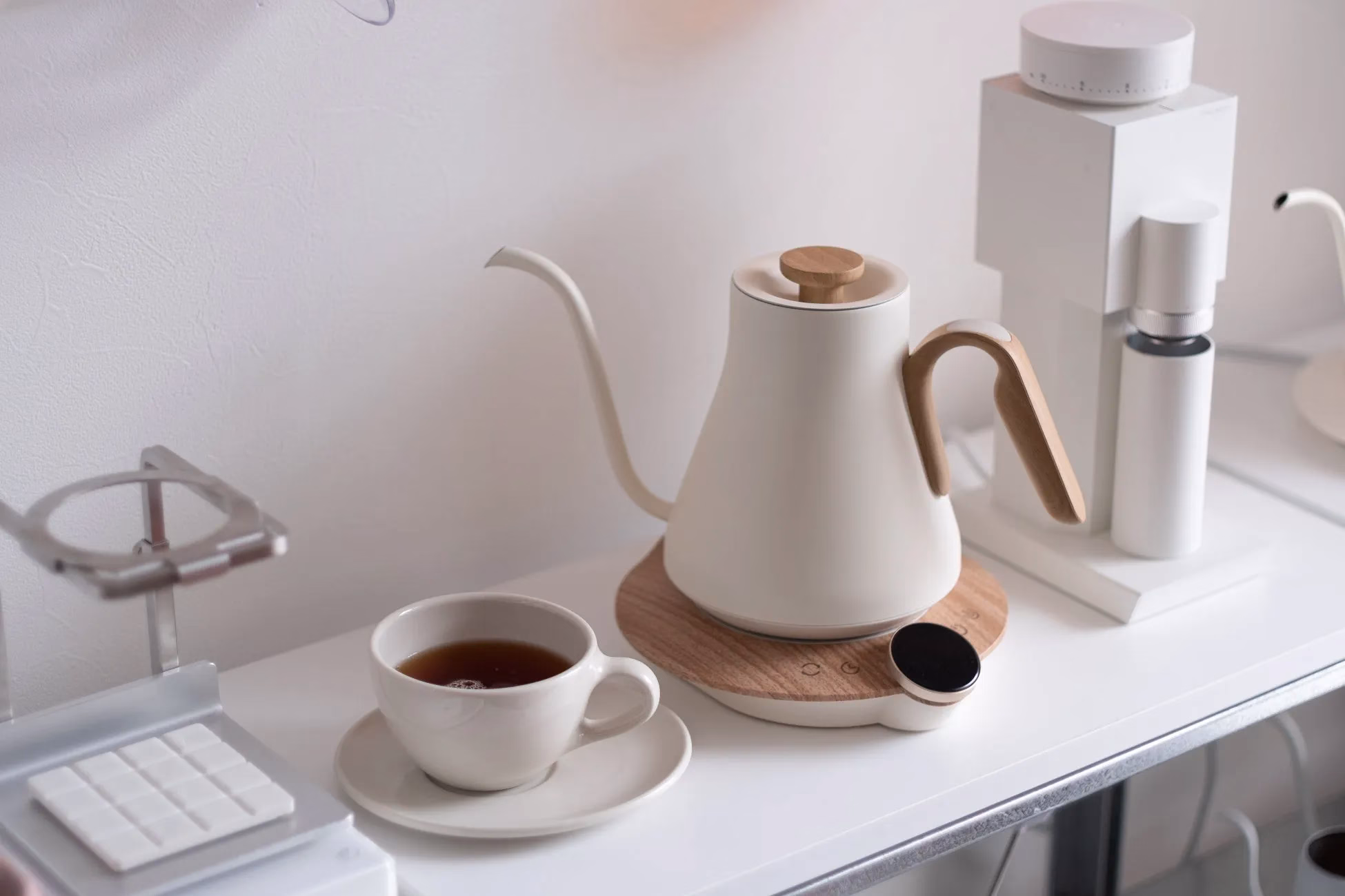 白い壁の前に置かれた、コーヒーポット、コーヒーミル、カップとソーサーに入ったお茶の静物写真。